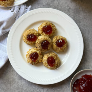 A white plate with seven Raspberry Thumbprint Cookies topped with red jam sits on a gray surface. Nearby are a spoonful of jam, more Jam Thumbprint Cookies on a plate, a glass of milk, and a white napkin.