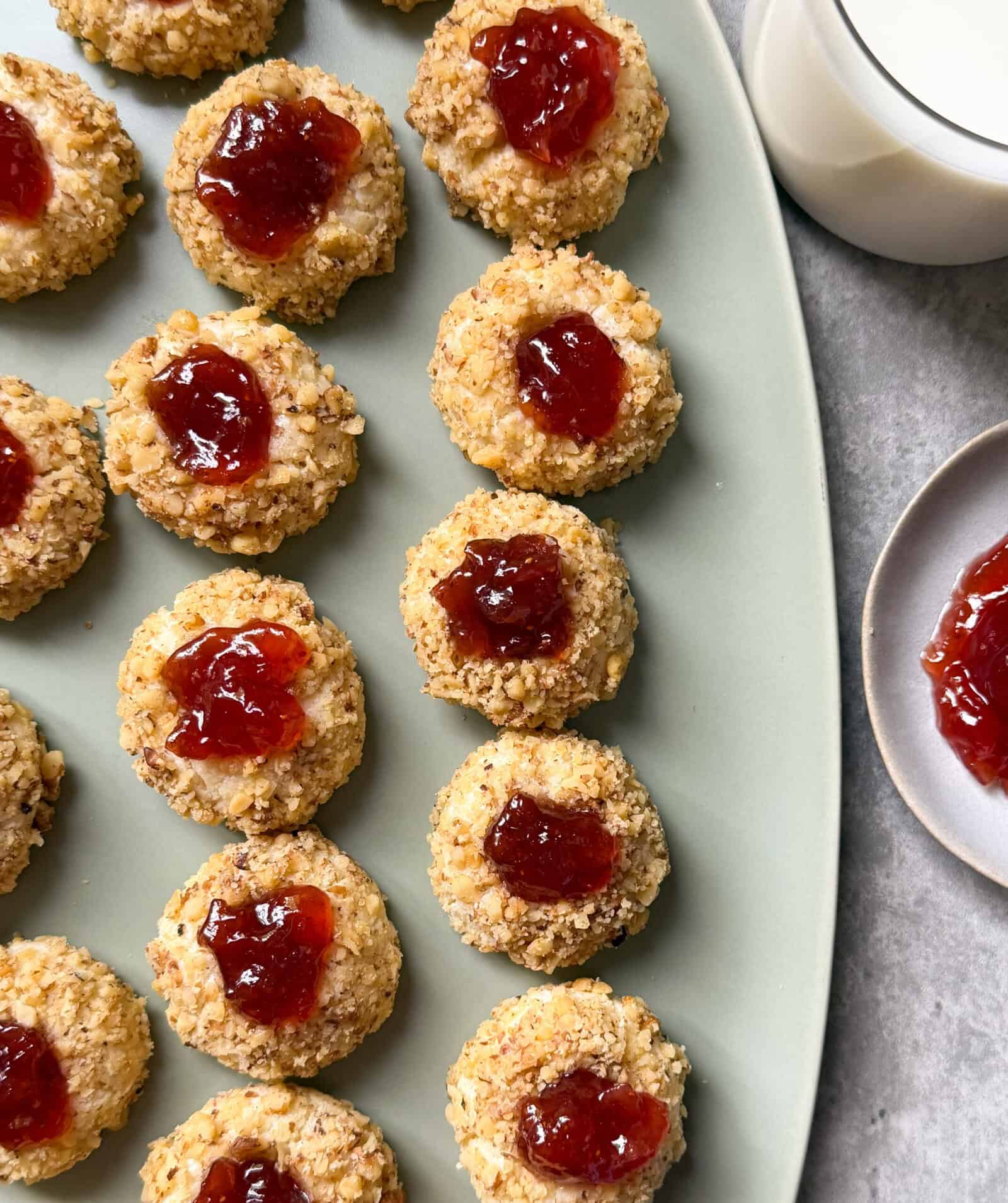 A platter of round Jam Thumbprint Cookies sits beside a glass of milk and a small plate with more cookies, all on a grey surface. The cookies are coated with chopped nuts and have a dollop of red jam in the center.