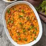 A baked casserole dish brimming with orange buffalo chicken dip, garnished with chopped green onions. Surrounding the tantalizing Buffalo Chicken Dip are tortilla chips and sliced carrots on a textured gray surface, inviting you to dive in and savor every bite.
