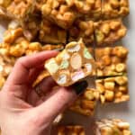 A hand holding a piece of no-bake dessert with colorful marshmallows and a caramel-like coating. Several pieces are scattered in the background on a white surface.