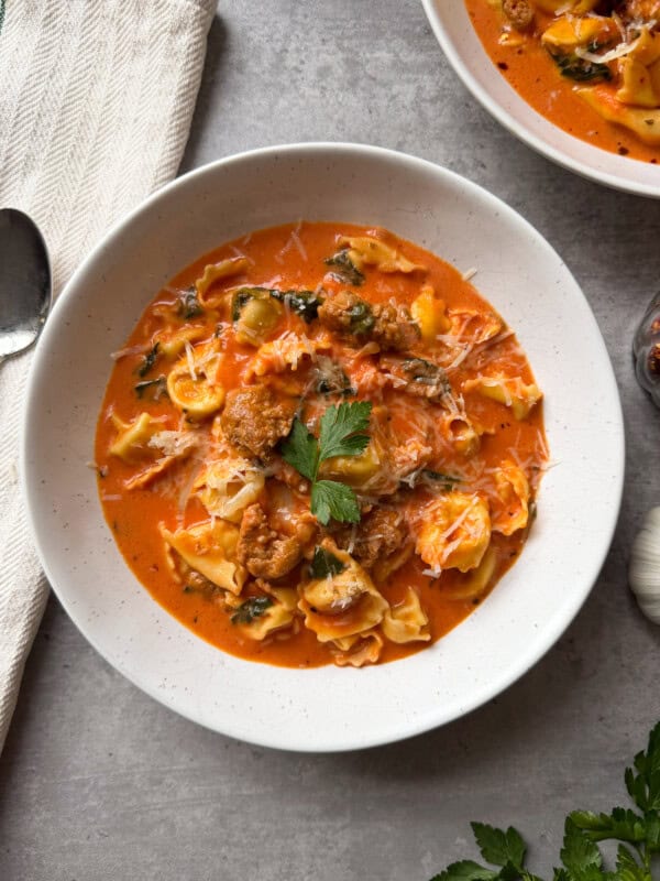 A white bowl filled with creamy Sausage Tortellini Soup topped with grated cheese and fresh parsley, placed on a gray surface near a spoon, napkin, and another partial bowl of soup.