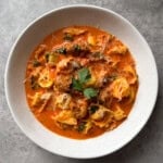 A white bowl filled with creamy sausage tortellini soup, topped with herbs and grated cheese, sits on a gray surface—an inviting bowl inspired by a classic sausage tortellini soup recipe.