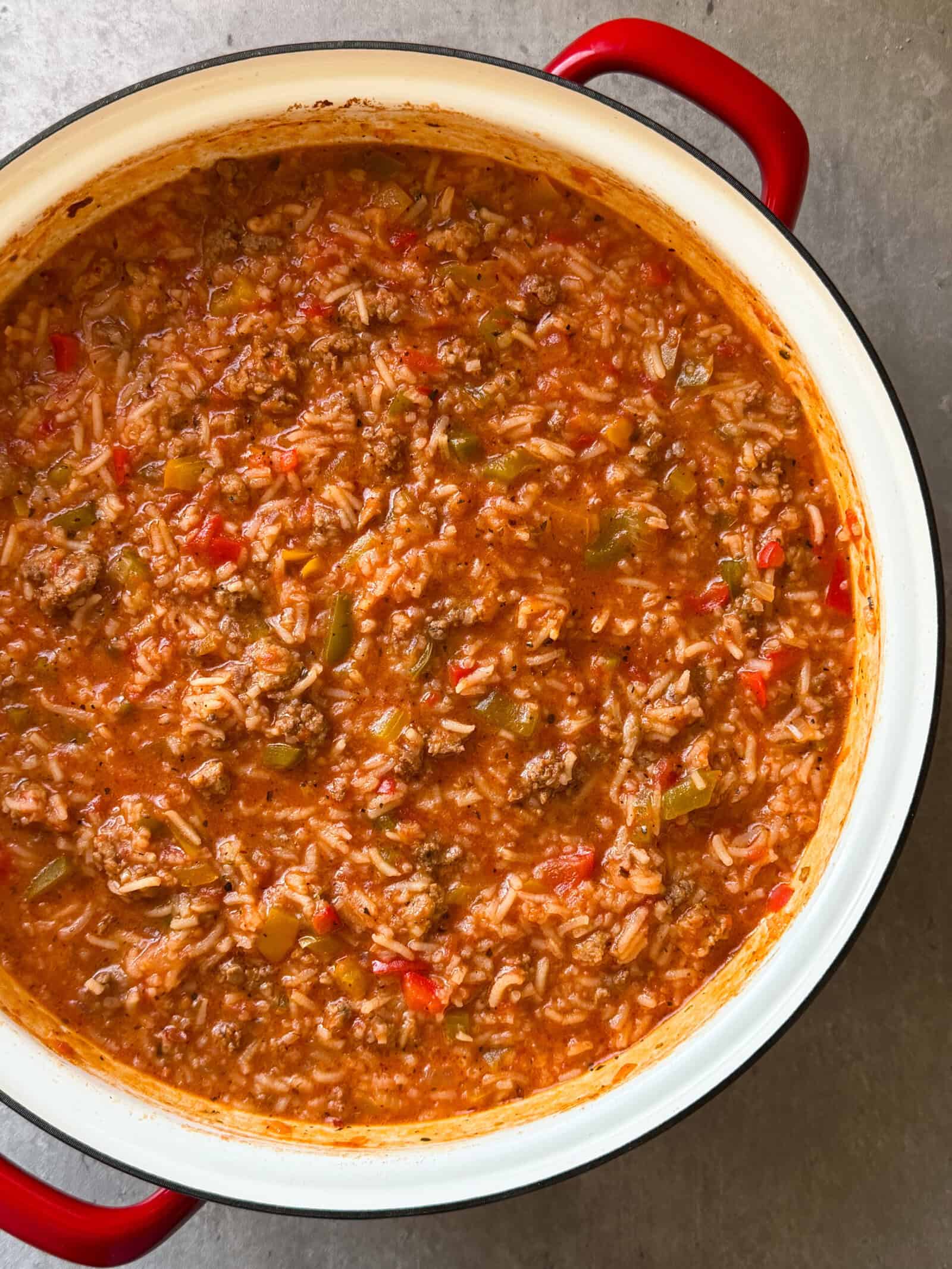 A large white pot with red handles contains a hearty Stuffed Pepper Soup Recipe made of ground beef, rice, tomatoes, and green peppers in a thick, red sauce. The freshly cooked dish is set on a gray surface.