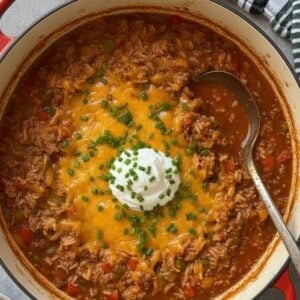 A pot of hearty chili, reminiscent of a Stuffed Pepper Soup recipe, topped with melted cheddar cheese, a dollop of sour cream, and chopped chives. A metal ladle rests inside, with a striped kitchen towel nearby.