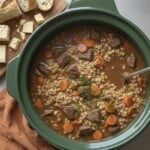 Slow cooker filled with crockpot beef barley soup, featuring chunks of beef, sliced carrots, and barley in rich broth. A wooden board holds sliced bread and cheese cubes. An orange cloth napkin sits underneath.