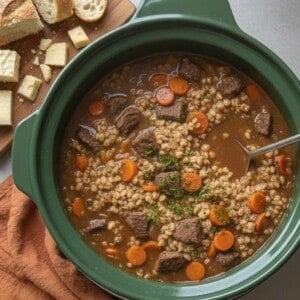 Slow cooker filled with crockpot beef barley soup, featuring chunks of beef, sliced carrots, and barley in rich broth. A wooden board holds sliced bread and cheese cubes. An orange cloth napkin sits underneath.