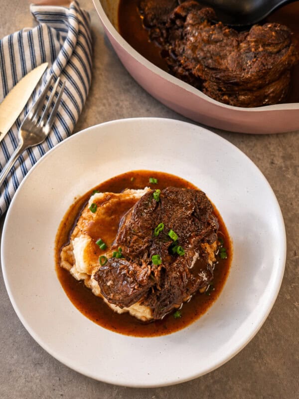 A plate of mashed potatoes topped with braised beef in gravy, garnished with chopped green onions, sits next to a cloth napkin and cutlery, with a pot of more beef in the background.