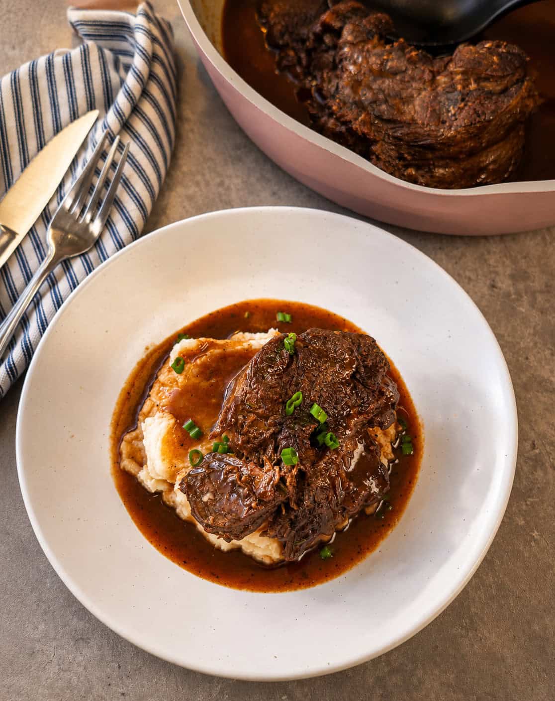 A plate of mashed potatoes topped with braised beef in gravy, garnished with chopped green onions, sits next to a cloth napkin and cutlery, with a pot of more beef in the background.