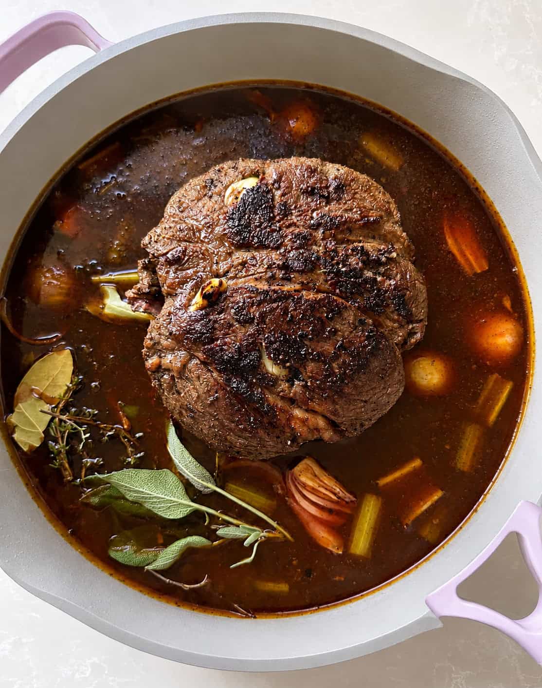 A browned pot roast sits in dark broth with carrots, celery, herbs, and garlic inside a light-colored pot with lavender handles.