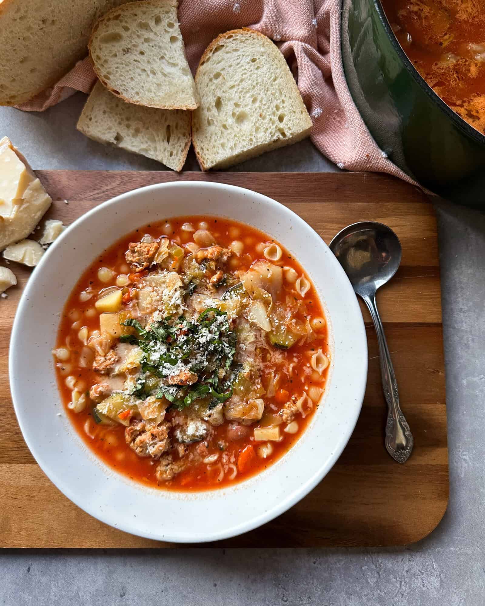 A bowl of hearty minestrone soup with pasta, sausage, and vegetables, topped with herbs and cheese, sits on a wooden board next to a spoon, sliced bread, and a block of cheese.