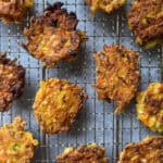 Golden brown vegetable fritters cooling on a metal rack lined with parchment paper. The fritters have a crispy texture and are arranged in a scattered pattern.