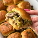 A hand holding a cheesy slider filled with green peppers, mushrooms, and beef, with more sliders visible on a white baking tray beneath. The sliders are topped with parsley.