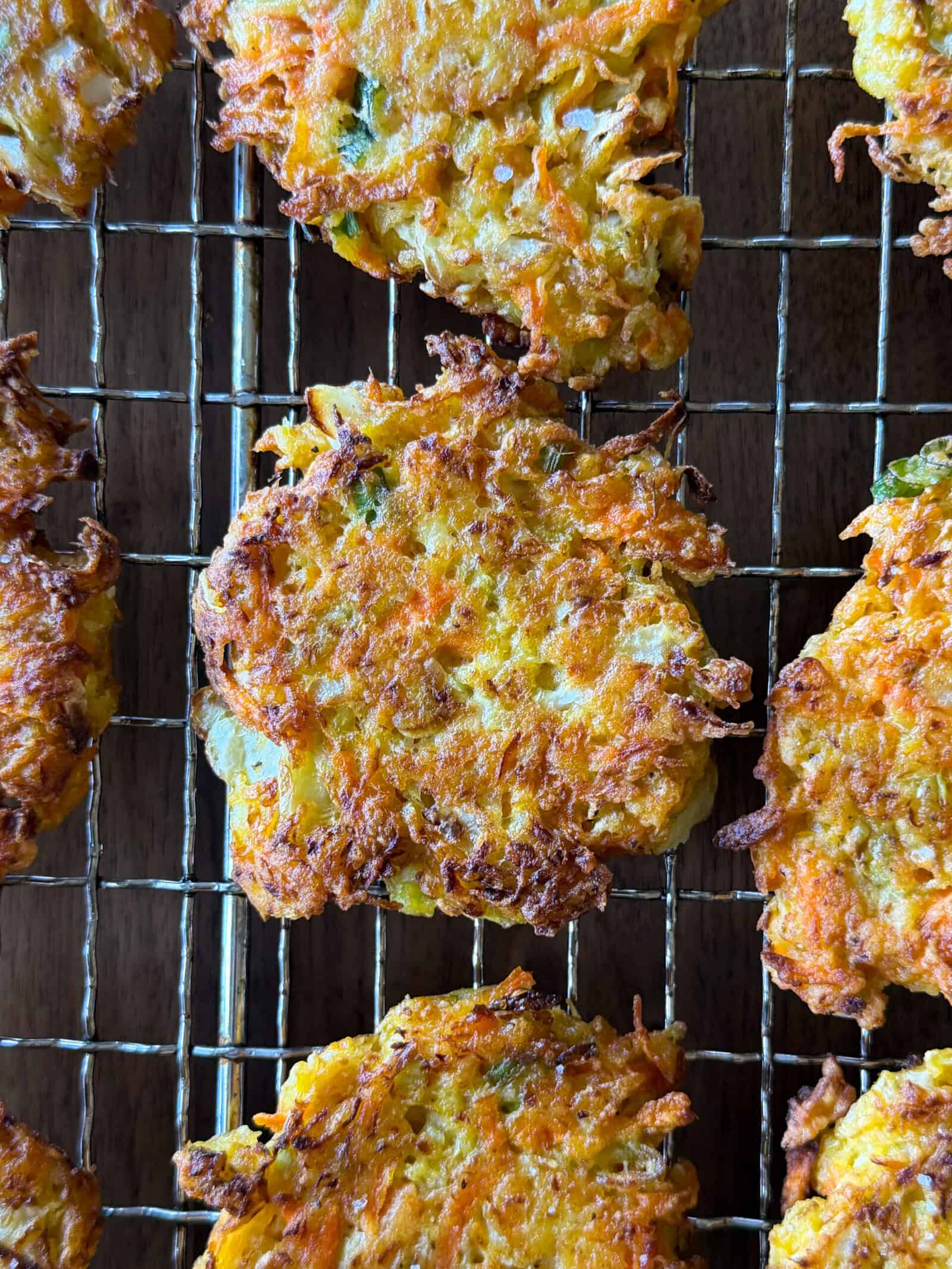 Several golden-brown cabbage fritters rest on a wire cooling rack, showing crisp, textured surfaces with visible shreds of carrot and herbs—a delicious highlight for anyone seeking an easy cabbage fritters recipe.