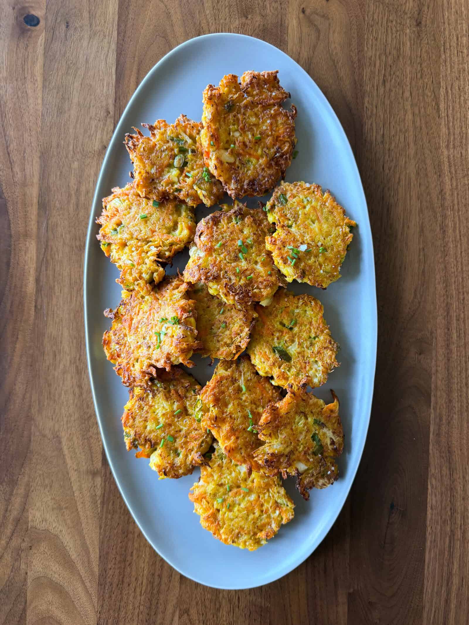 Oval plate with a pile of golden-brown, crispy cabbage fritters on a wooden table. The fritters have visible pieces of green zucchini, orange carrot, and shredded cabbage, making them the perfect choice for anyone looking for an easy cabbage fritters recipe.