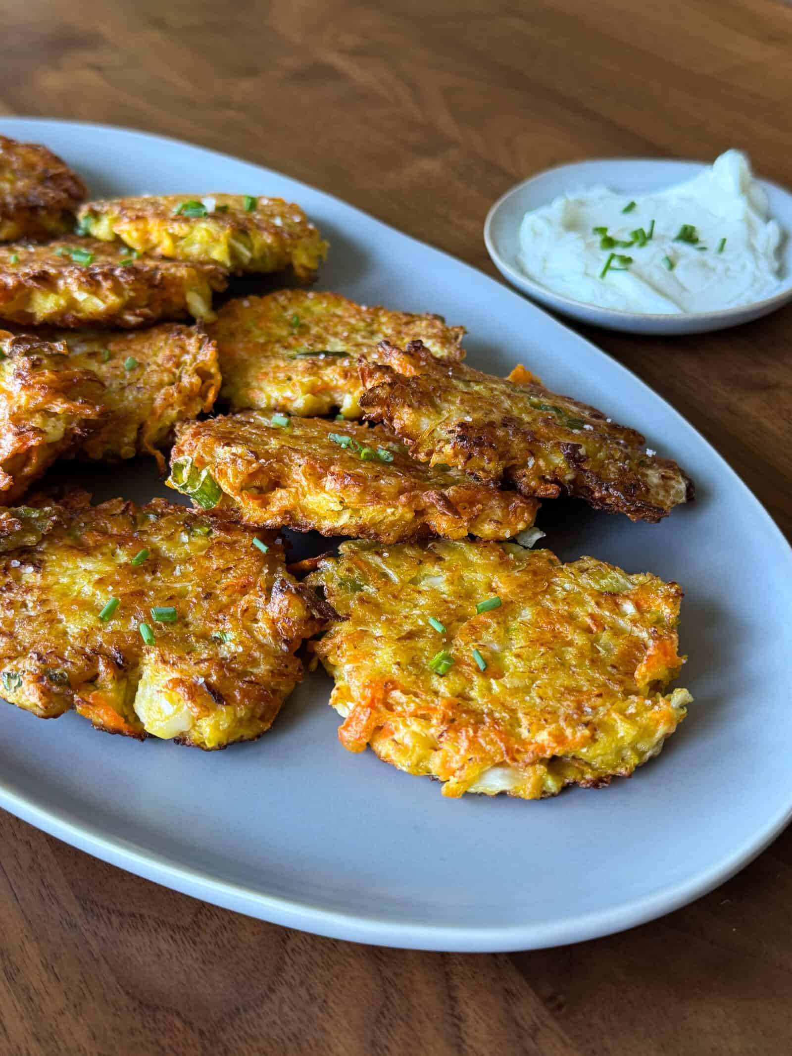 A gray plate holds several golden-brown cabbage fritters, garnished with chopped herbs. A small dish of creamy white dipping sauce with green garnish sits nearby on a wooden table, making this an inviting display for anyone seeking easy cabbage recipes.