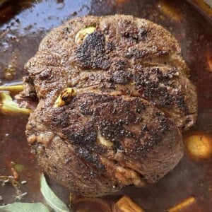 A browned roast beef sits in rich, dark broth with visible garlic cloves, herbs, celery, and onions, all in a pot. The surface of the meat is seared and slightly crispy.
