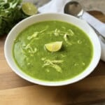 A white bowl of Green Goddess Soup with shredded chicken, garnished with a lime wedge. A spoon, napkin, fresh cilantro, and a halved lime are nearby on a wooden board—perfect for fans of healthy soup recipes.