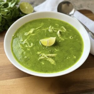 A white bowl of Green Goddess Soup with shredded chicken, garnished with a lime wedge. A spoon, napkin, fresh cilantro, and a halved lime are nearby on a wooden board—perfect for fans of healthy soup recipes.