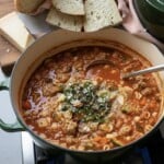 A pot of hearty Minestrone simmers on the stove, filled with vegetables, pasta, and ground meat, then topped with herbs and grated cheese. Slices of rustic bread and cheese rest on a cutting board nearby.