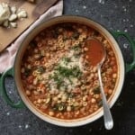 A pot of hearty Minestrone Soup with small pasta, vegetables, and ground meat, topped with grated cheese and herbs, sits on a dark countertop. Nearby, sliced bread and cheese await, complementing this classic Minestrone recipe.