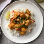 A plate of white rice topped with golden chicken pieces drizzled in orange sauce. Garnished with chopped green onions and a wedge of lime on the side, set on a gray surface with a striped cloth nearby.