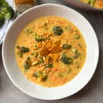 A bowl of creamy broccoli and cheese soup garnished with tortilla chips, surrounded by small pieces of bread. The soup contains visible broccoli florets and shredded carrots, creating a colorful and appetizing appearance.