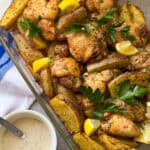 A glass baking dish filled with roasted chicken thighs and potato wedges, garnished with lemon slices and fresh parsley. A small bowl of creamy sauce with herbs sits beside the dish on a blue and white striped cloth.