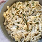 A pan filled with mafaldine pasta in a creamy white sauce, garnished with grated cheese and chopped herbs. The wavy pasta is mixed with small pieces of mushrooms. The dish is presented on a light countertop.