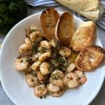 A white plate filled with cooked shrimp garnished with herbs and garlic, accompanied by toasted baguette slices. A loaf of bread and a bunch of fresh parsley are in the background on a striped cloth.