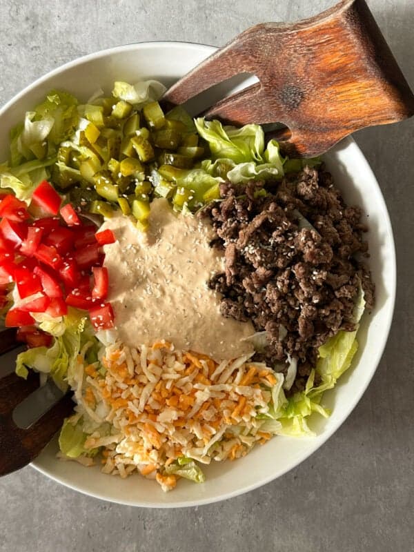 A bowl of Big Mac salad contains ground beef, shredded cheese, chopped tomatoes, diced pickles, lettuce, and creamy dressing, with wooden salad tongs resting on the edge. Try this Big Mac salad recipe for a delicious twist on a classic!.