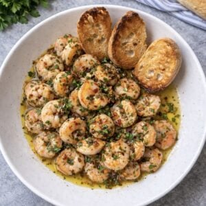 A white bowl filled with shrimp cooked in a herbed garlic butter sauce, inspired by gamberi alla sambuca, garnished with chopped parsley and served with three toasted baguette slices. Fresh parsley and a baguette are in the background.