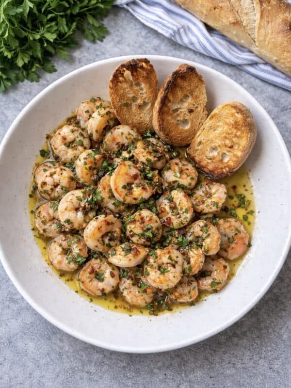 A white bowl filled with shrimp cooked in a herbed garlic butter sauce, inspired by gamberi alla sambuca, garnished with chopped parsley and served with three toasted baguette slices. Fresh parsley and a baguette are in the background.
