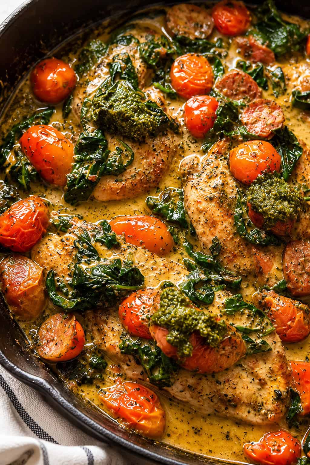 Chicken breasts in a creamy sauce with kale, cherry tomatoes, and dollops of green pesto, cooked in a black skillet.