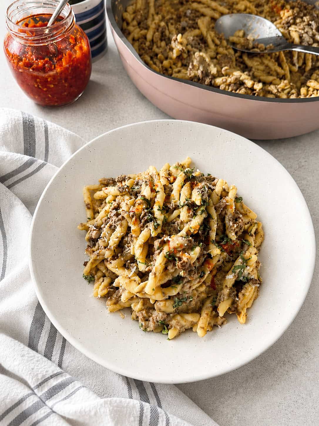 A plate of pasta with a creamy sauce and herbs sits on a white surface beside a striped towel, a jar of red sauce, and a skillet with more pasta and a serving spoon.
