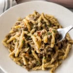 A close-up of a plate of pasta with ground meat sauce, garnished with herbs. A fork holds a bite of the pasta above the dish. A striped napkin and part of a red sauce jar are visible in the background.