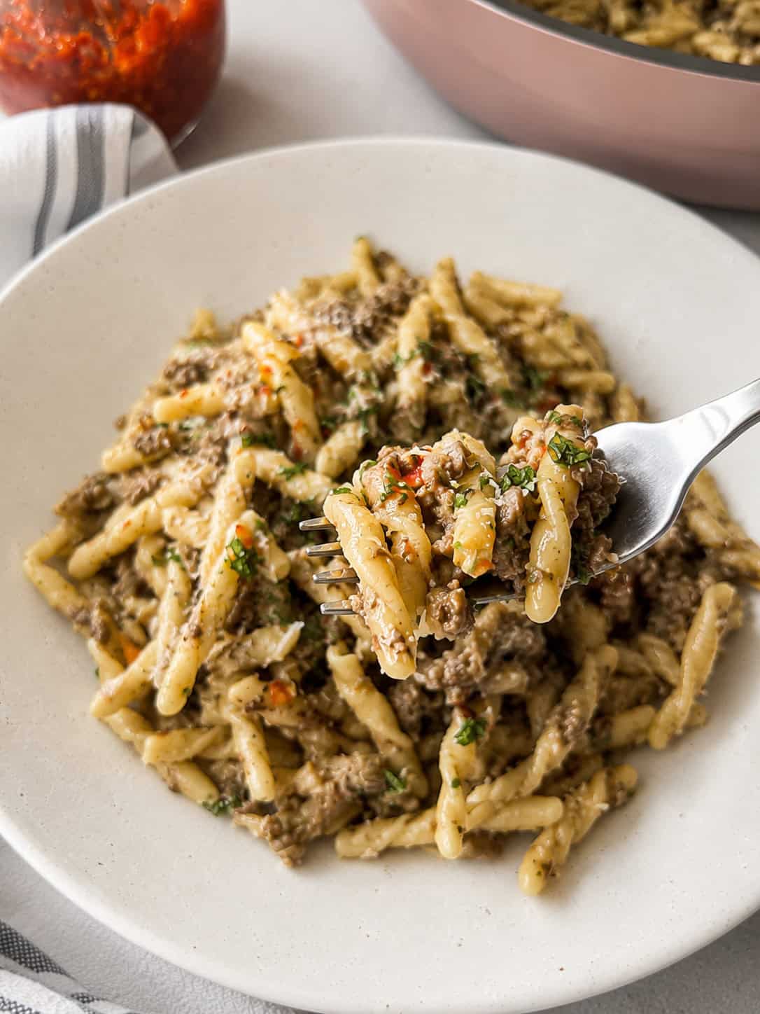 A close-up of a plate of pasta with ground meat sauce, garnished with herbs. A fork holds a bite of the pasta above the dish. A striped napkin and part of a red sauce jar are visible in the background.