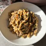 A bowl of pasta topped with a creamy sauce, ground beef, and finely chopped vegetables, garnished with fresh herbs. The dish is served in a white bowl on a table, partially lit by sunlight.