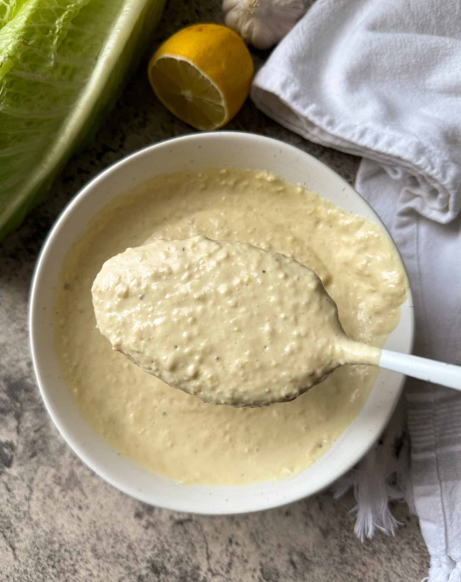 A spoon holds up a thick, creamy white sauce above a bowl&mdash;homemade Caesar dressing recipe&mdash;surrounded by halved lemon, leafy lettuce, and a white kitchen towel on a gray countertop.