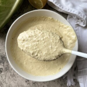 A spoonful of creamy, textured homemade caesar dressing recipe is held over a white bowl filled with more of the same mixture. A lemon half, lettuce, and a white cloth are nearby on a gray surface.