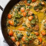 A skillet filled with a creamy dish featuring chicken breasts, cherry tomatoes, and spinach, topped with dollops of green pesto sauce. The ingredients are cooked in a rich, golden sauce.