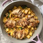 A skillet filled with pappardelle pasta, spinach, and sliced grilled chicken. The pasta is creamy and seasoned with herbs, and the chicken is golden brown with visible grill marks. A white cloth is partially visible in the background.