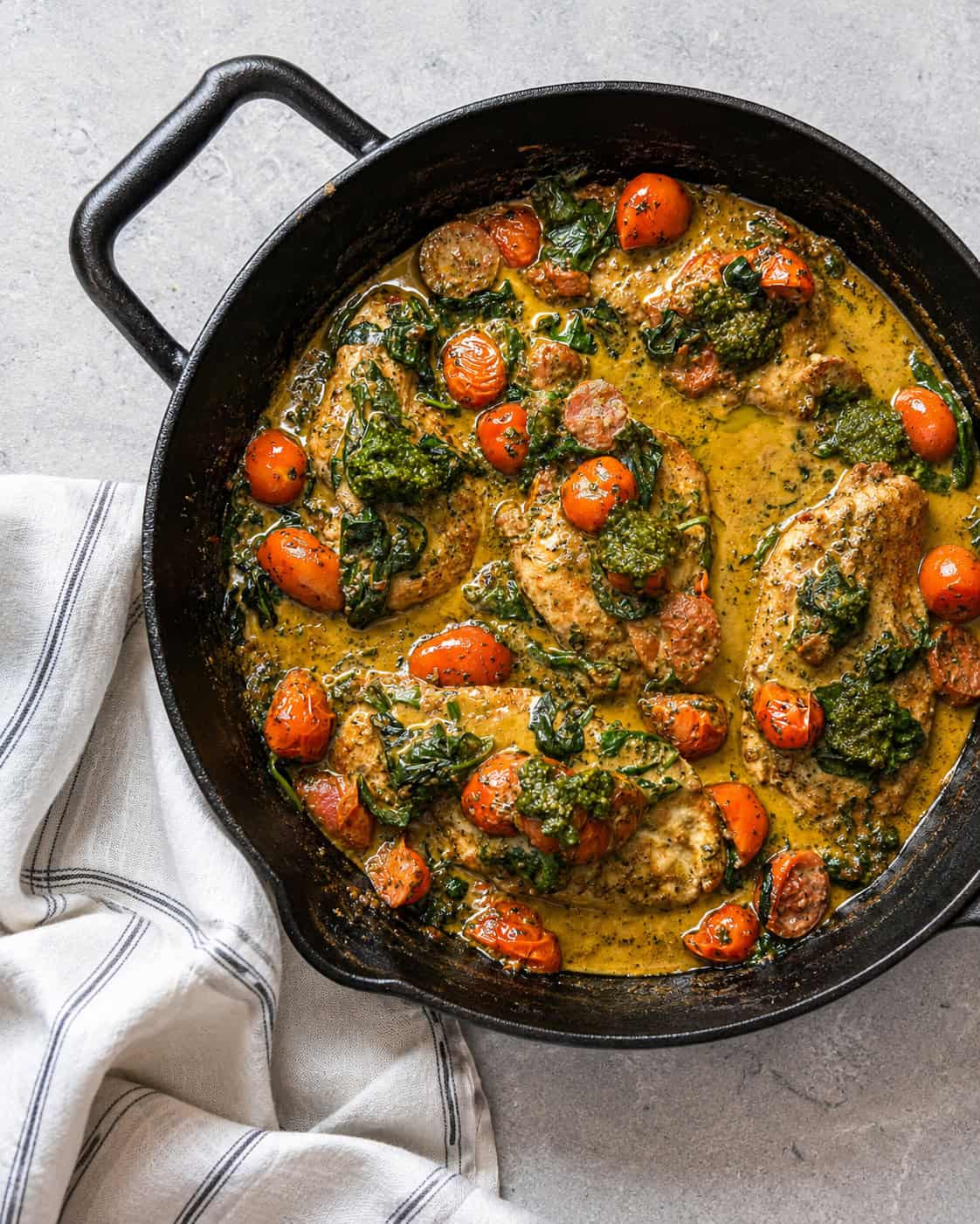 A black skillet filled with chicken breasts, cherry tomatoes, spinach, and a green herb sauce sits on a gray countertop beside a folded white-striped kitchen towel.