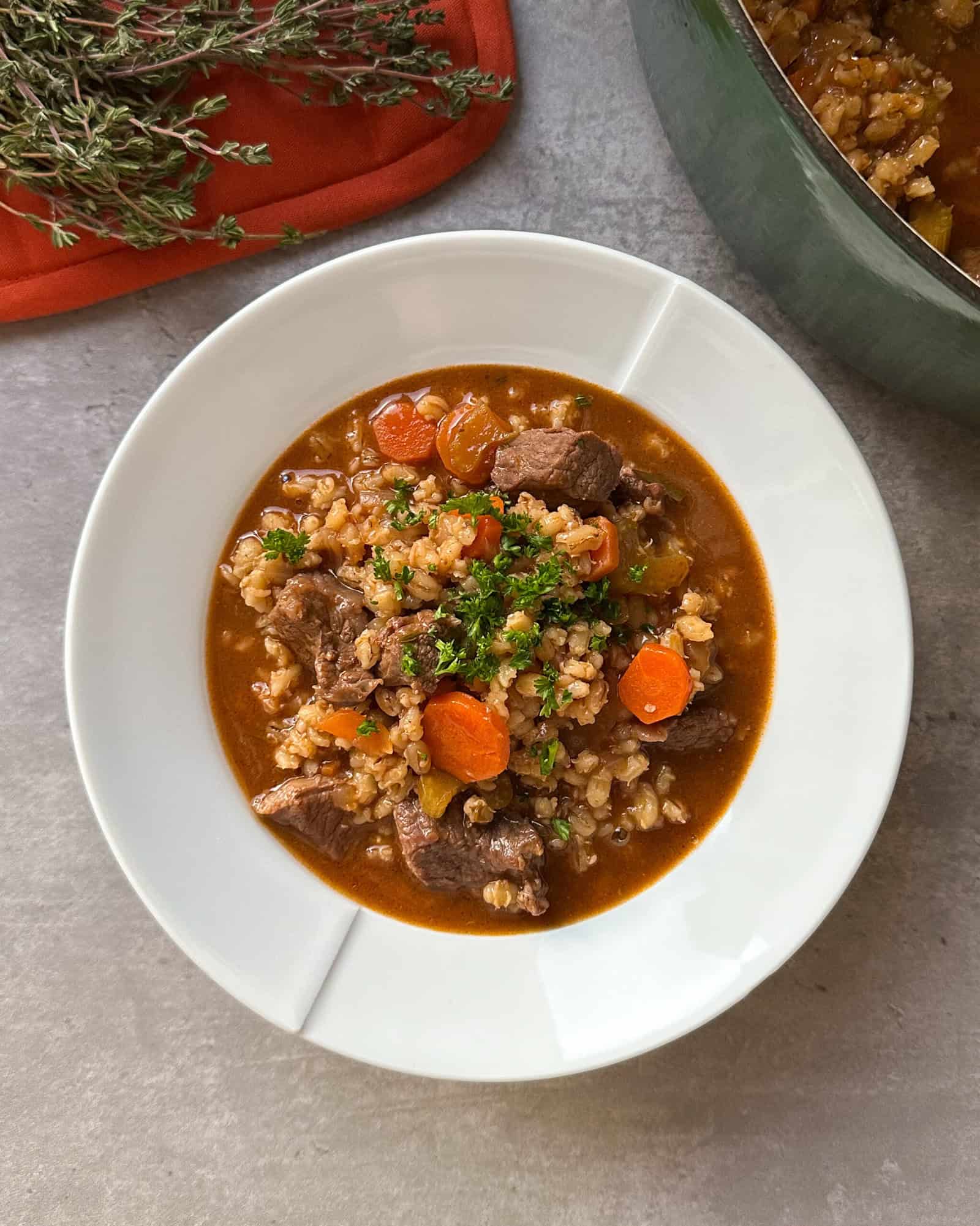 A white bowl filled with beef and barley stew, featuring chunks of beef, sliced carrots, barley, and garnished with fresh herbs—the perfect crockpot beef barley soup. A pot of stew and fresh thyme are visible in the background.