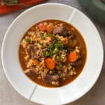 A white plate filled with beef stew, featuring chunks of beef, carrots, and pearl barley in a rich brown sauce. The dish is garnished with chopped parsley, set on a gray table with a glimpse of a green pot in the background.