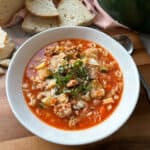 A white bowl of hearty soup sits on a wooden table, filled with chunks of vegetables, pasta, and sausage in a tomato-based broth. It is garnished with fresh herbs and served alongside slices of crusty bread. A spoon rests nearby.