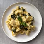 A plate of cheese tortellini mixed with creamy sauce, wilted spinach, and topped with fresh basil leaves, served on a round white plate. The background is a textured gray surface.