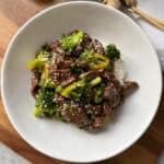 A white bowl filled with stir-fried beef and broccoli served over white rice. The dish is garnished with sesame seeds and appears to be on a wooden surface with chopsticks and a piece of ginger nearby.