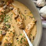 Creamy garlic chicken with mushrooms is in a pan. The dish is garnished with chopped herbs. A white spoon rests in the sauce. Garlic bulbs and fresh thyme are placed beside the pan on a striped cloth.