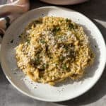 A white plate with creamy orzo pasta garnished with grated cheese and chopped herbs. The pasta is evenly spread and appears rich and textured, with a hint of pepper on top. Sunlight casts a warm glow on the dish, enhancing its appetizing look.
