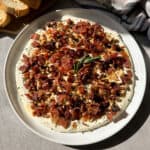 A round dish topped with a creamy spread, crumbled bacon, and nuts, garnished with a sprig of rosemary. Slices of bread are in the background on the left. The dish is placed on a gray surface.
